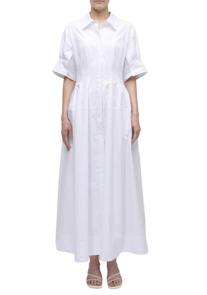 A person wearing the Simkhai Mollie Shirt Dress—a long, white, button-down dress with short sleeves and a collar—stands in white heeled sandals against a plain white background.