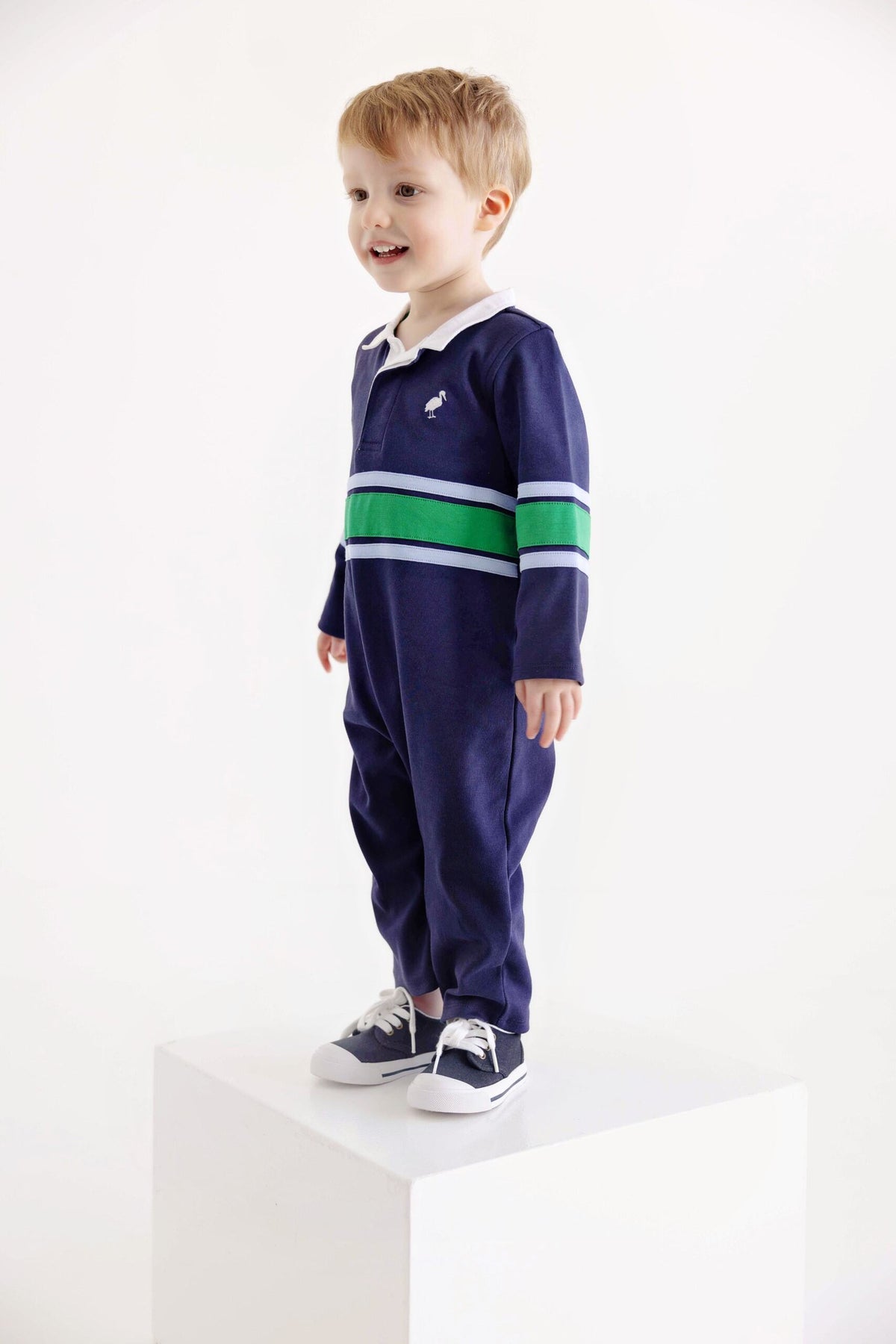 A young child wearing The Beaufort Bonnet Company’s Baby Boys’ Sir Proper’s Rugby Romper—navy blue with green and white stripes—stands smiling on a white pedestal against a plain white background.