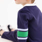 A young child with light hair sits sideways, dressed in The Beaufort Bonnet Company’s Baby Boys’ Sir Proper’s Rugby Romper—a navy long-sleeve piece crafted from soft Pima cotton, featuring light blue and green stripes on the arms. The romper is paired with matching navy pants, set against a clean white background.