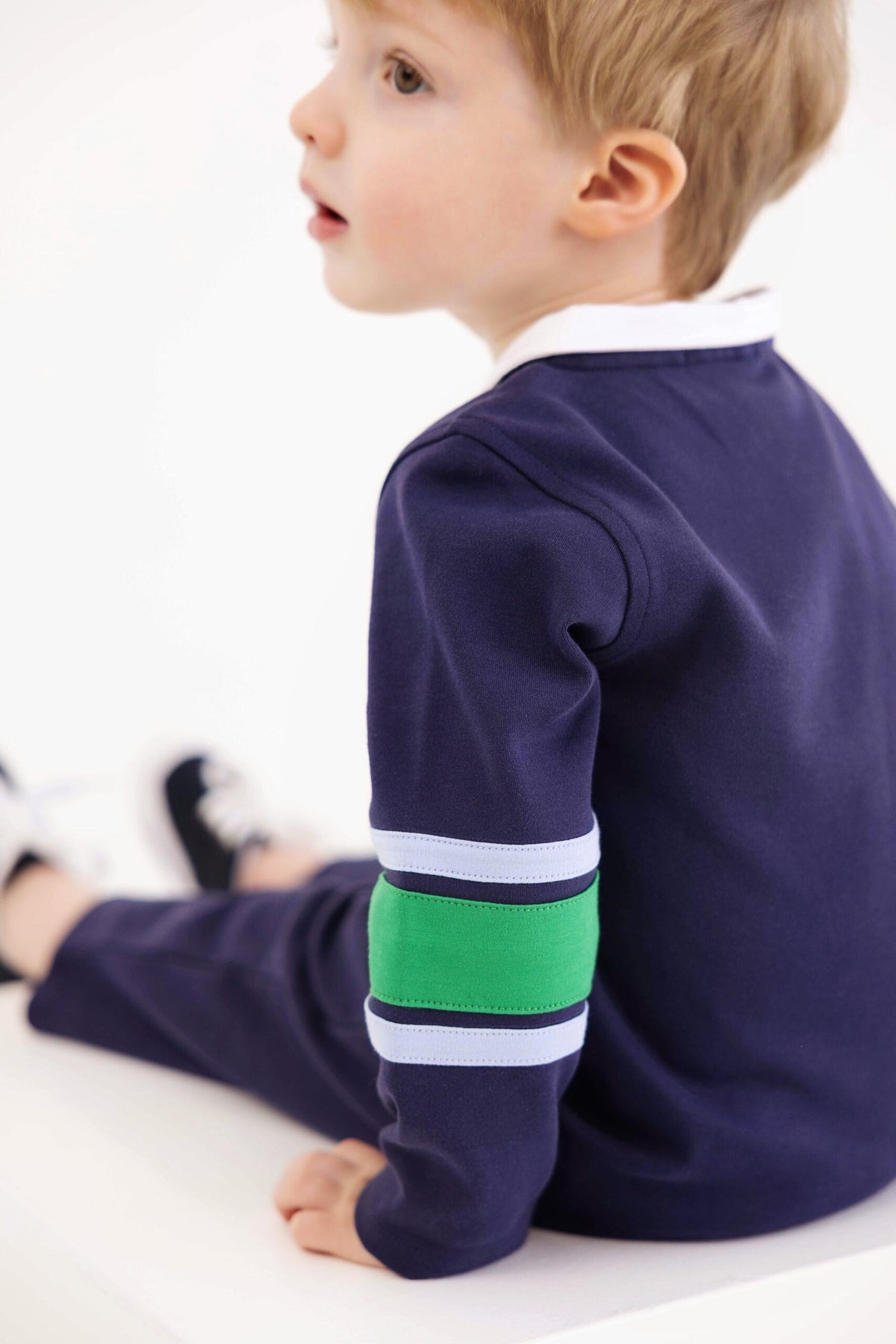 A young child with light hair sits sideways, dressed in The Beaufort Bonnet Company’s Baby Boys’ Sir Proper’s Rugby Romper—a navy long-sleeve piece crafted from soft Pima cotton, featuring light blue and green stripes on the arms. The romper is paired with matching navy pants, set against a clean white background.