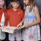 Three children stand together reading a book; one girl, wearing the Baby Girls’ and Girls’ Sleeveless Cindy Lou Sash Dress by The Beaufort Bonnet Company in a Kensington Court Plaid, holds the book open while a boy and another girl look at the pages. They are dressed in coordinated plaid and solid-colored outfits.