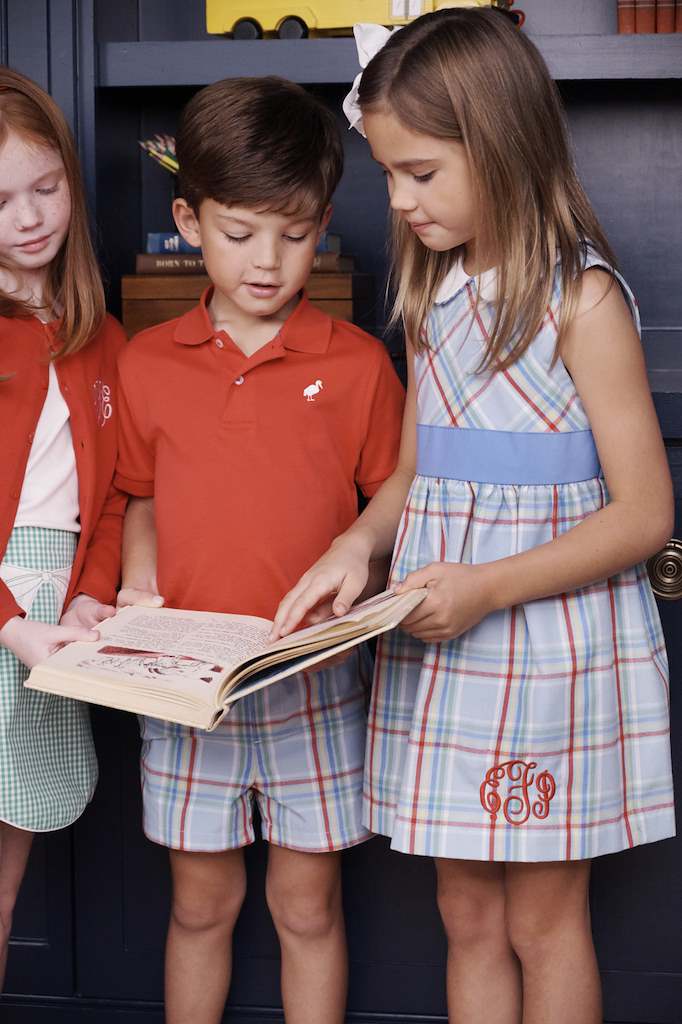 Three children stand together reading a book; one girl, wearing the Baby Girls’ and Girls’ Sleeveless Cindy Lou Sash Dress by The Beaufort Bonnet Company in a Kensington Court Plaid, holds the book open while a boy and another girl look at the pages. They are dressed in coordinated plaid and solid-colored outfits.