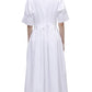A woman stands facing away, wearing the Simkhai Mollie Shirt Dress—a long, white, short-sleeved dress with a fitted waist and collar—paired with light-colored heels.