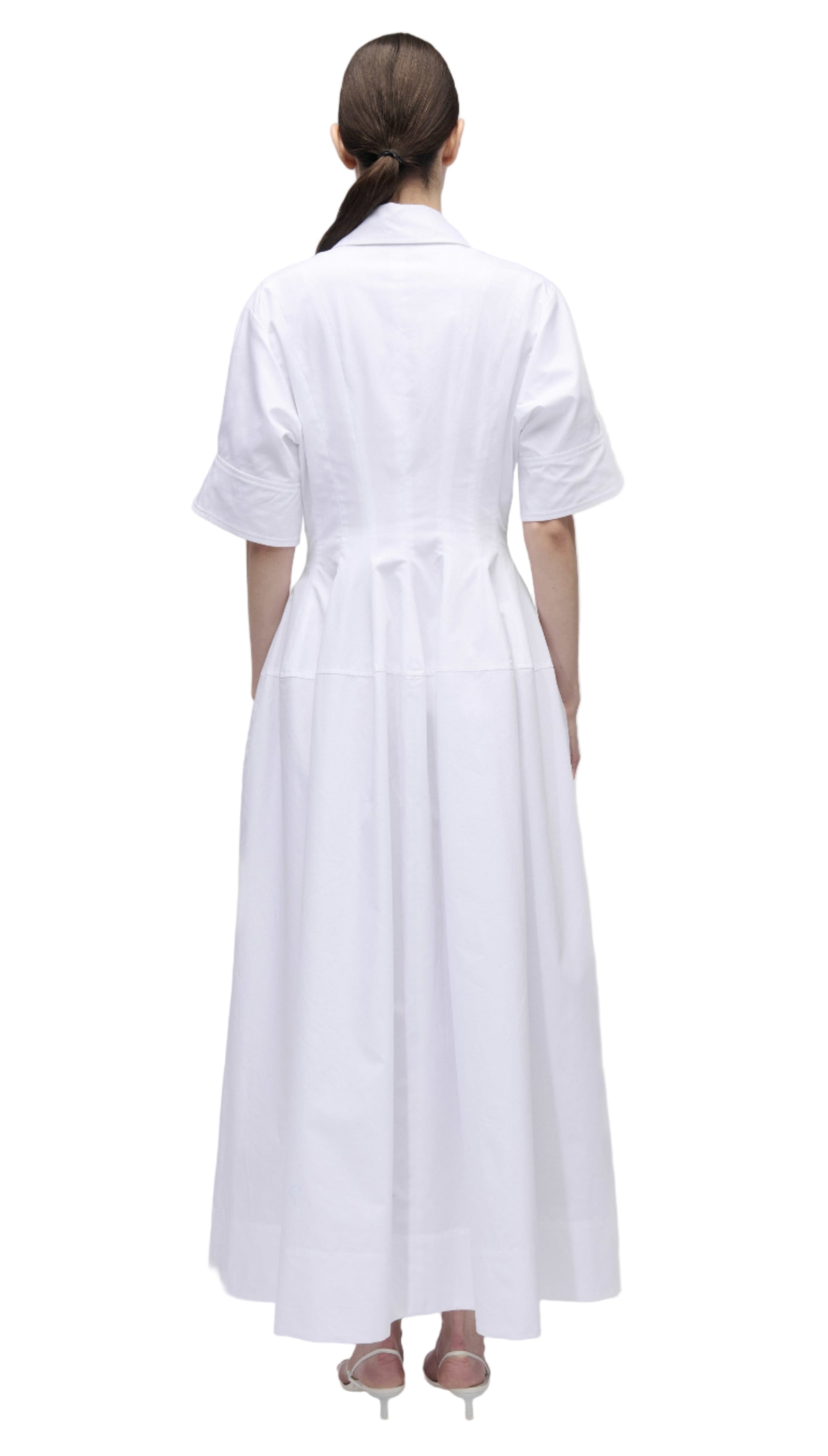 A woman stands facing away, wearing the Simkhai Mollie Shirt Dress—a long, white, short-sleeved dress with a fitted waist and collar—paired with light-colored heels.
