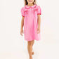 A young girl stands on a white background wearing The Beaufort Bonnet Company Baby Girls’ and Girls’ Tabitha’s Teacher’s Pet Dress in Hamptons Hot Pink, which features puff sleeves, white buttons, and charming red ribbon bows.