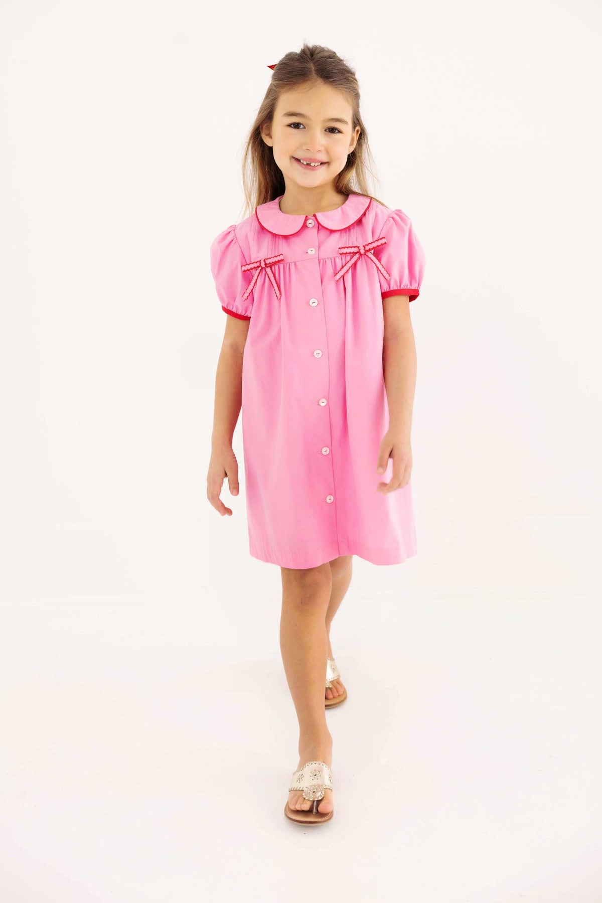 A young girl stands on a white background wearing The Beaufort Bonnet Company Baby Girls’ and Girls’ Tabitha’s Teacher’s Pet Dress in Hamptons Hot Pink, which features puff sleeves, white buttons, and charming red ribbon bows.