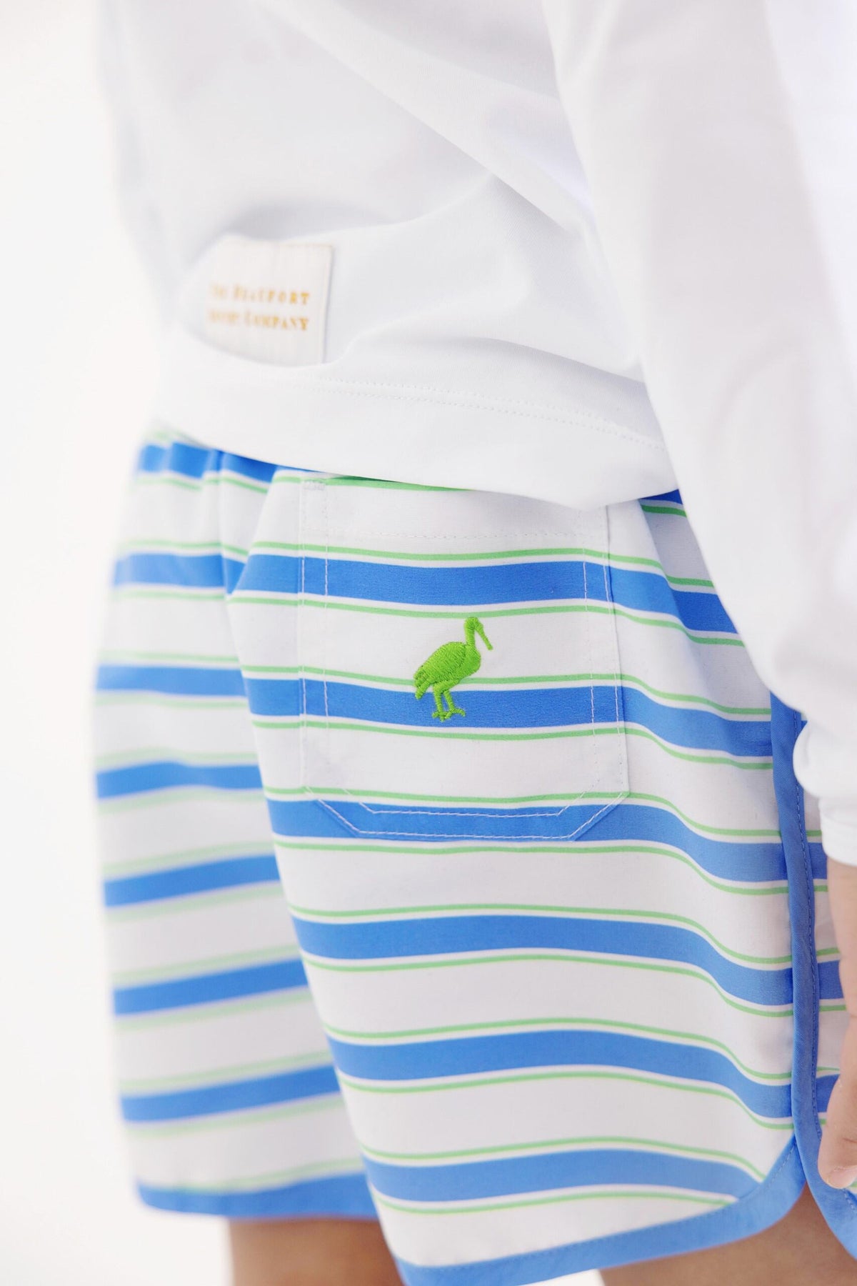 A close-up of a person wearing The Beaufort Bonnet Company’s Baby Boys’ and Boys’ Tanner Trunks in the Seabrook Stripe pattern—blue, green, and white striped shorts featuring a green bird embroidered on the back pocket.