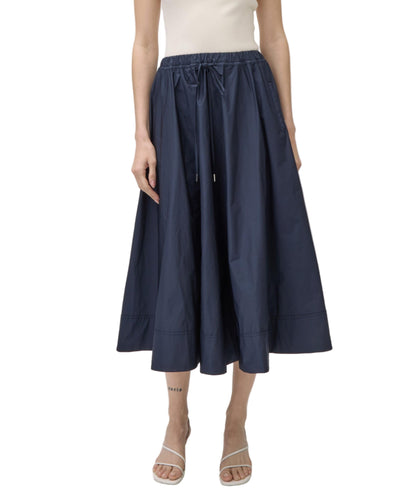 A person wearing a white sleeveless top, the Simkhai Flynn Skirt—a navy blue, high-waisted midi A-line skirt with an elastic waistband—and white sandals.