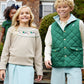Two children wearing light blue and Saratoga Gate Green outfits, including the Baby Boys’ and Boys’ Vaughn Vest by The Beaufort Bonnet Company, walk outside together smiling, while another child stands in the background near a house.