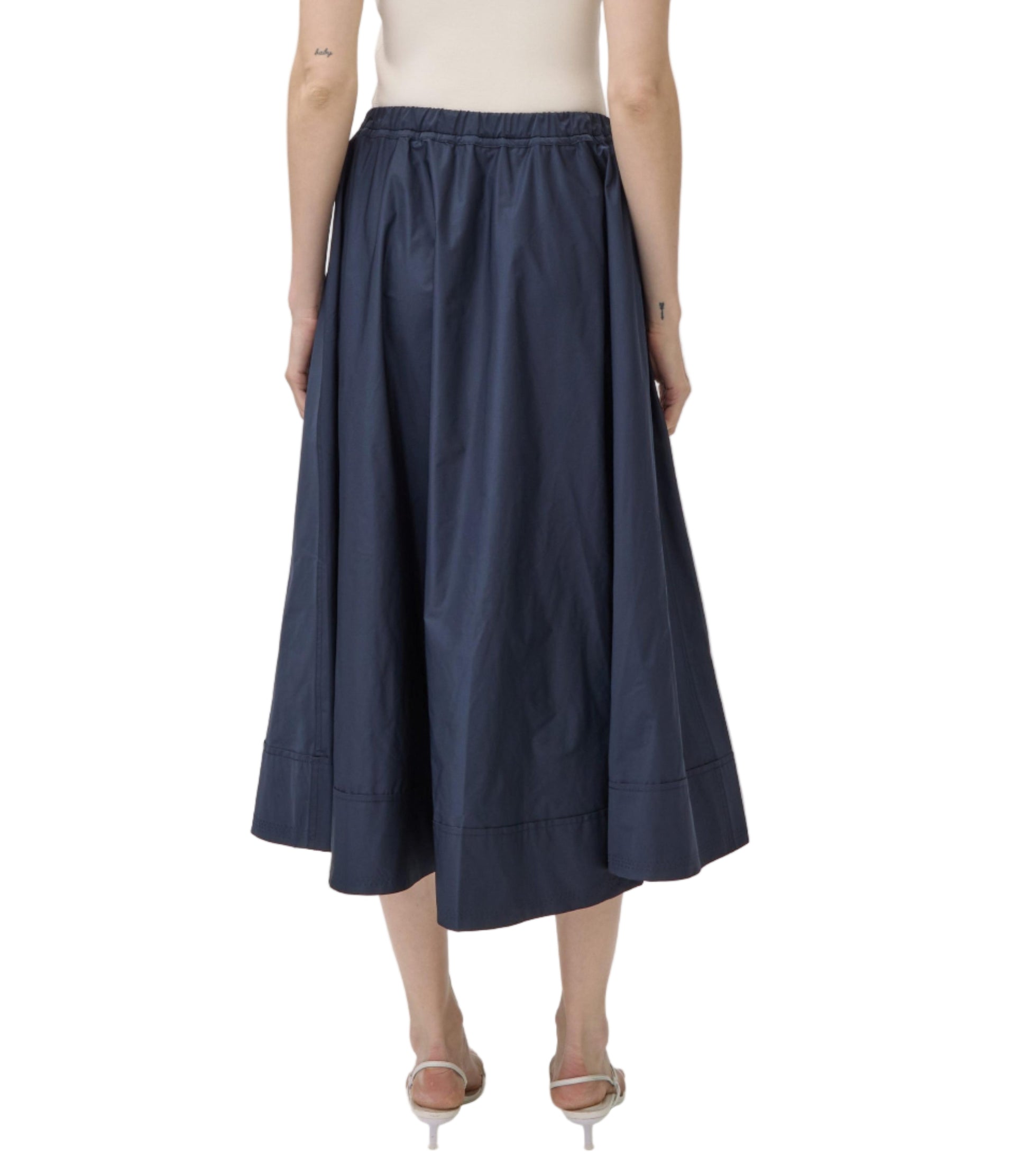 A person wearing the Simkhai Flynn Skirt in navy blue, ankle-length, paired with heeled sandals, shown from the back against a white background.