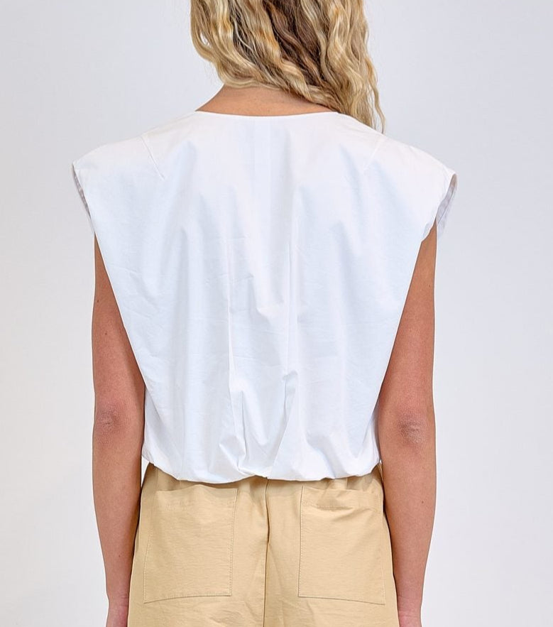 A person stands with their back to the camera, wearing the Simkhai Wrenli Pleated Top in white and beige high-waisted shorts against a plain background.