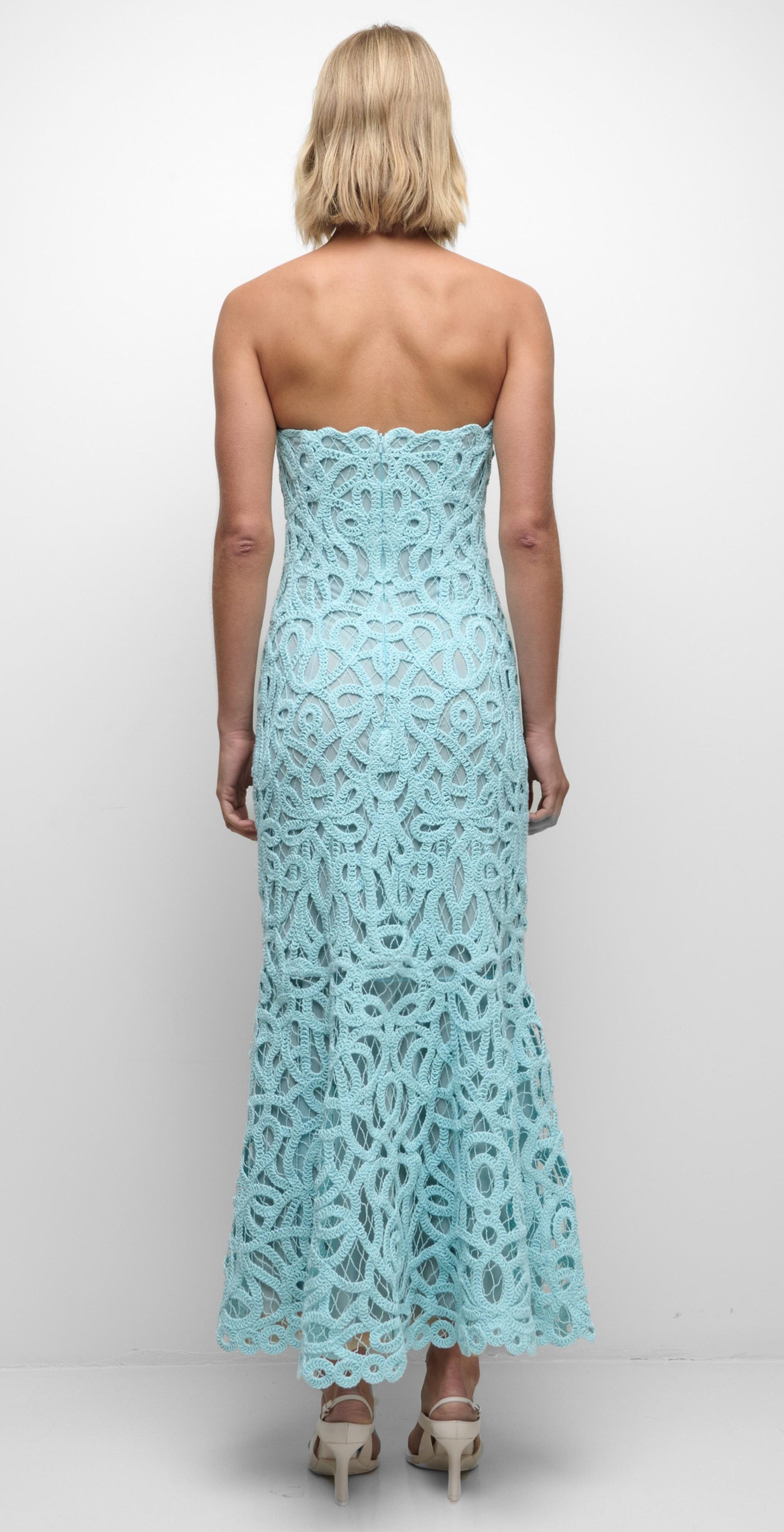 A woman facing away wears the Simkhai Clairis Lace Bustier Midi Dress in light blue, featuring a strapless bodice and flared hem, styled with beige heeled sandals.