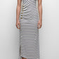 A woman wears the Simkhai Becker Cotton Jersey Midi Dress—sleeveless, white with navy horizontal stripes and side button details—paired with white sandals, standing against a plain background.