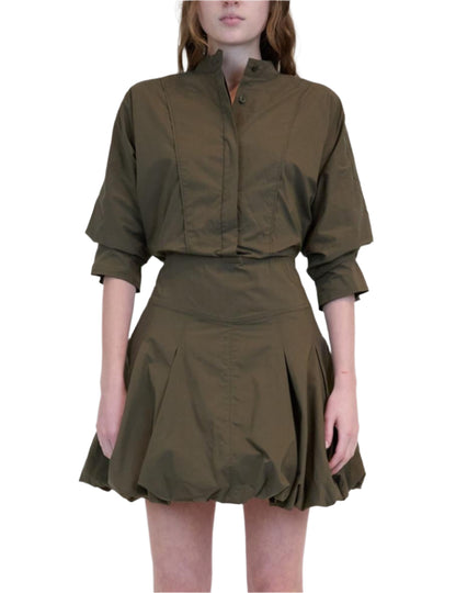 A person wearing the Effie Bubble Mini Dress by Simkhai—an olive green cotton poplin dress with a fitted waist, button-up top, and flared balloon skirt—stands against a white background.