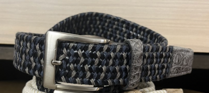 A close-up of the W. Kleinberg Men's Solid Stretch Belt with Croc Tabs in woven blue and gray, featuring a silver buckle and partially wrapped in protective plastic.