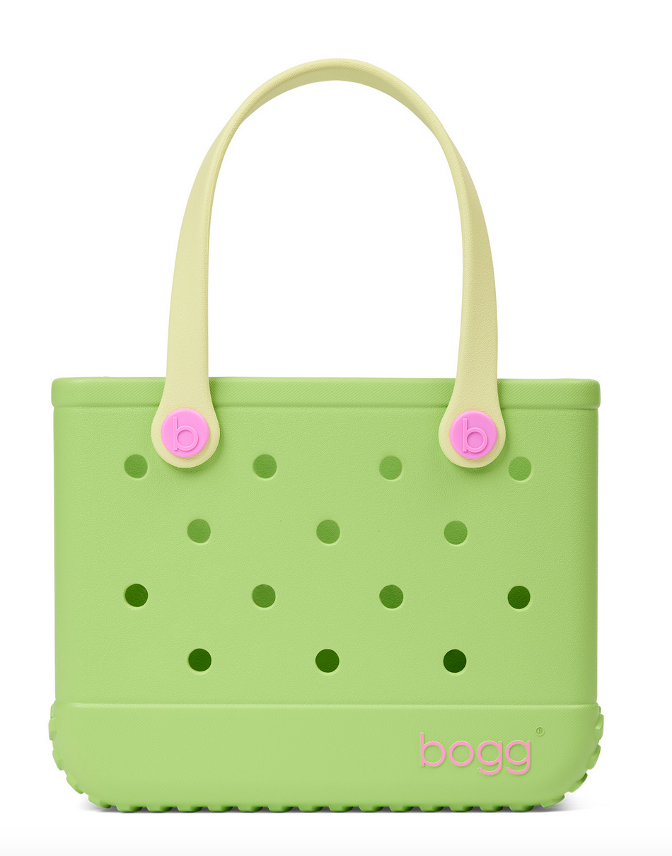 Light green Bitty Bogg® Bag by Bogg Bags, a compact tote featuring cream handles, round holes throughout, pink button accents, and a pink “bogg” logo at the bottom right corner. This washable bag is perfect for everyday use.