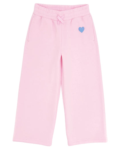 The Girls’ Cozy Sweatpant by Feather 4 Arrow features a light pink, wide-leg design with an elastic waistband, front pockets, flared bottoms, and a small blue heart detail on the left side.