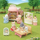 Several Calico Critters animal figurines enjoy pastries and drinks at an outdoor patisserie scene, featuring the Village Cake Shop Starter Set with its charming pink cart, delicious sponge cakes adorned with intricate decorations, all set in a park-like area complete with grass, trees, and a wooden fence.