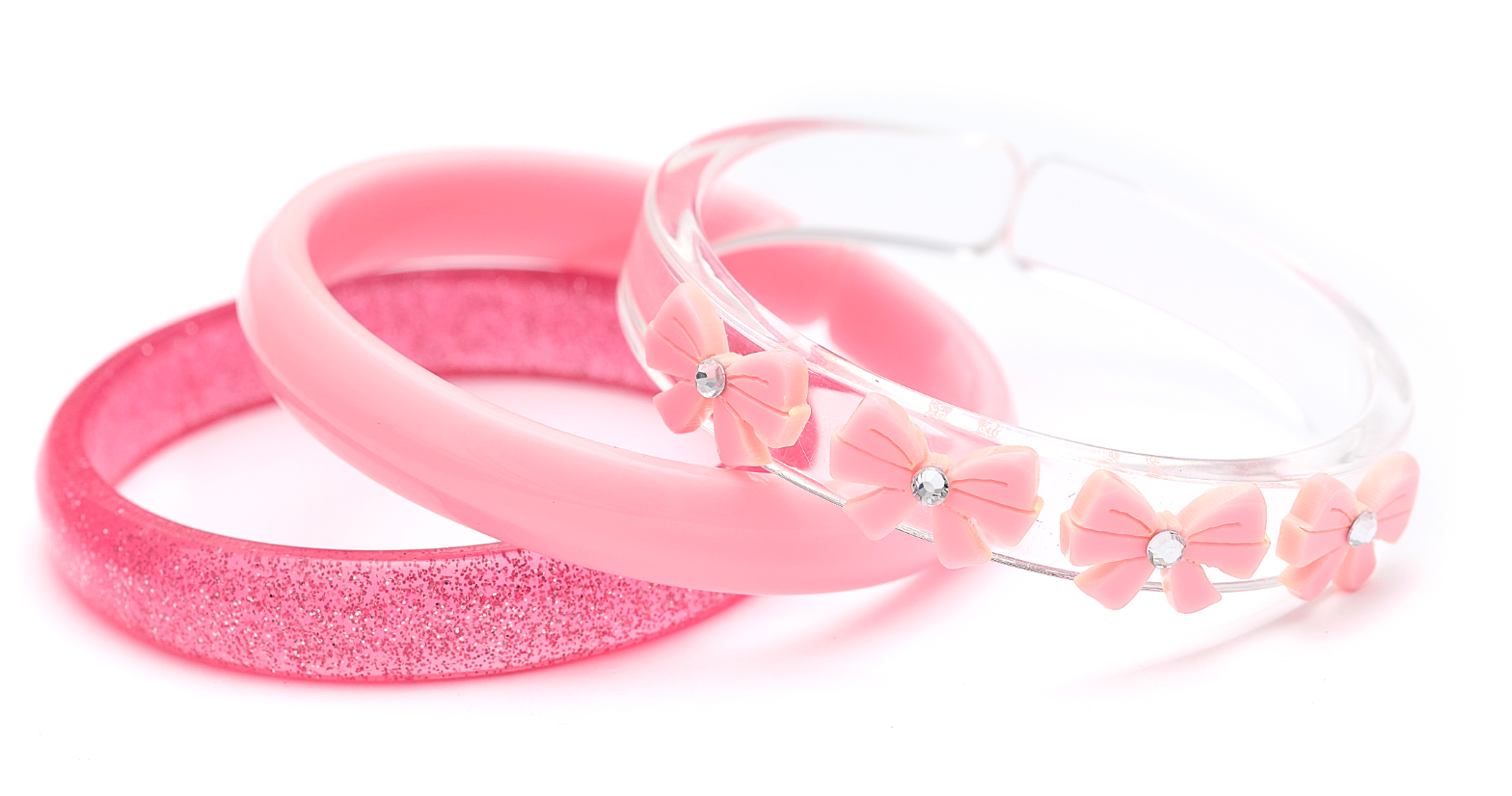 Three pink bracelets from Lillies & Roses’ Bows Vintage Pink Bangles set—featuring one glittery bangle, one solid pink bangle, and one clear bracelet embellished with pink bows and rhinestones—arranged on a white background.