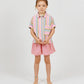 A young girl stands barefoot against a plain background, wearing Arsene & Les Pipelettes' Girls' Vichy Short in pink gingham seersucker, a colorful striped shirt, and a matching hair tie, with a beaded anklet on her left ankle.