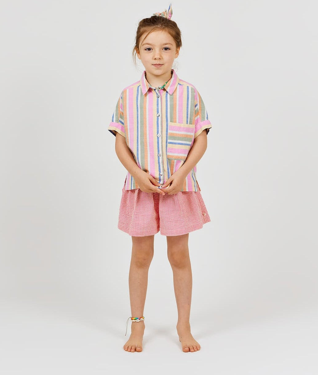 A young girl stands barefoot against a plain background, wearing Arsene & Les Pipelettes' Girls' Vichy Short in pink gingham seersucker, a colorful striped shirt, and a matching hair tie, with a beaded anklet on her left ankle.