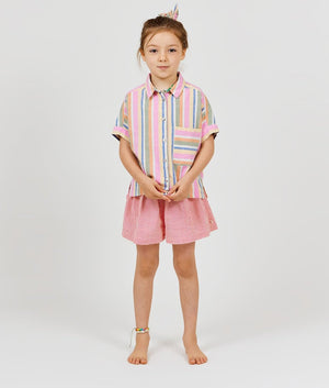A young girl stands barefoot against a plain background, wearing Arsene & Les Pipelettes' Girls' Vichy Short in pink gingham seersucker, a colorful striped shirt, and a matching hair tie, with a beaded anklet on her left ankle.