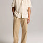 A person stands against a plain background wearing the vince Cove Stripe SS Cabana, a short-sleeve beige cotton-hemp shirt with a double-stripe pattern, paired with tan wide-leg trousers and brown shoes.