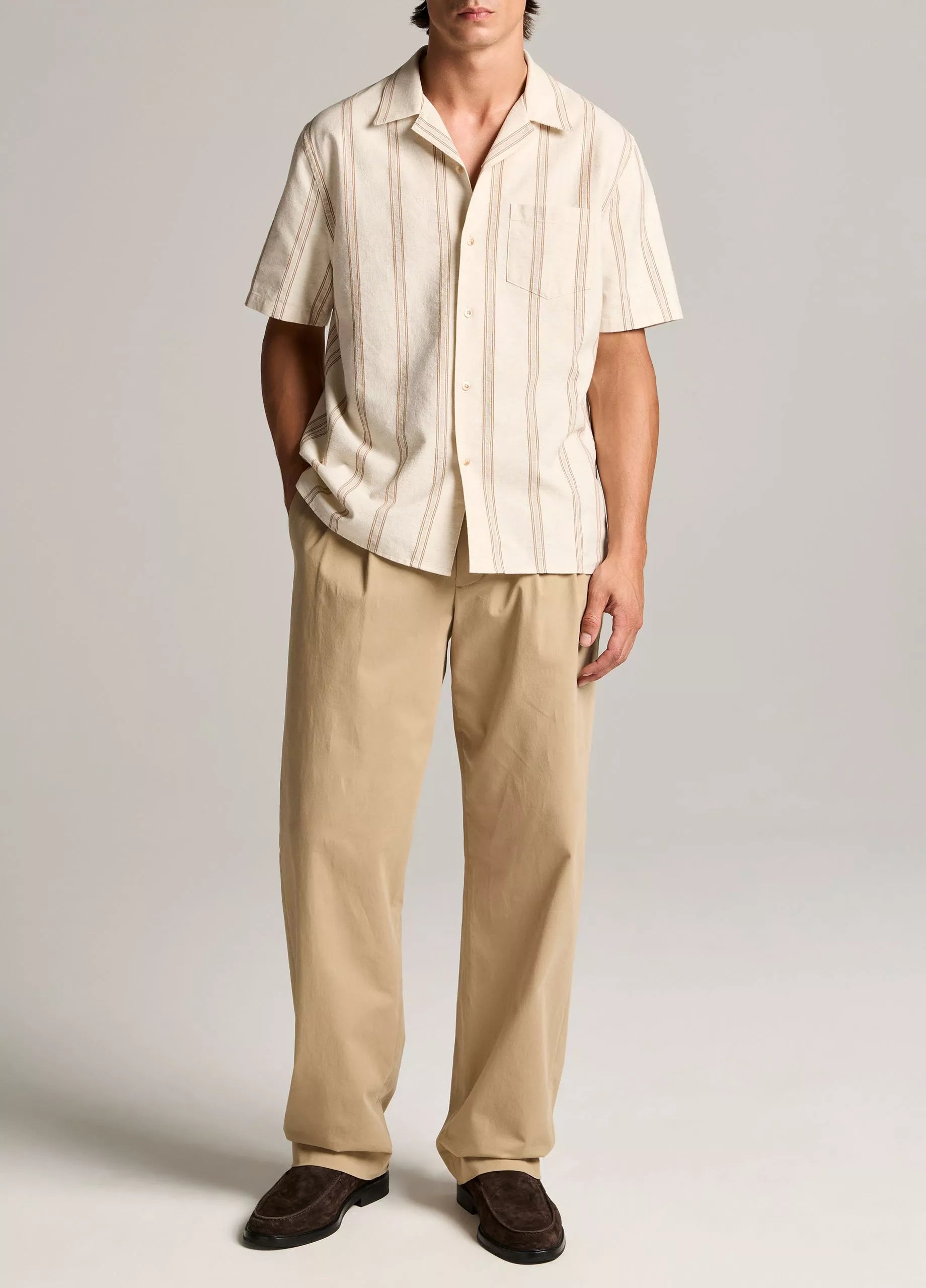 A person stands against a plain background wearing the vince Cove Stripe SS Cabana, a short-sleeve beige cotton-hemp shirt with a double-stripe pattern, paired with tan wide-leg trousers and brown shoes.