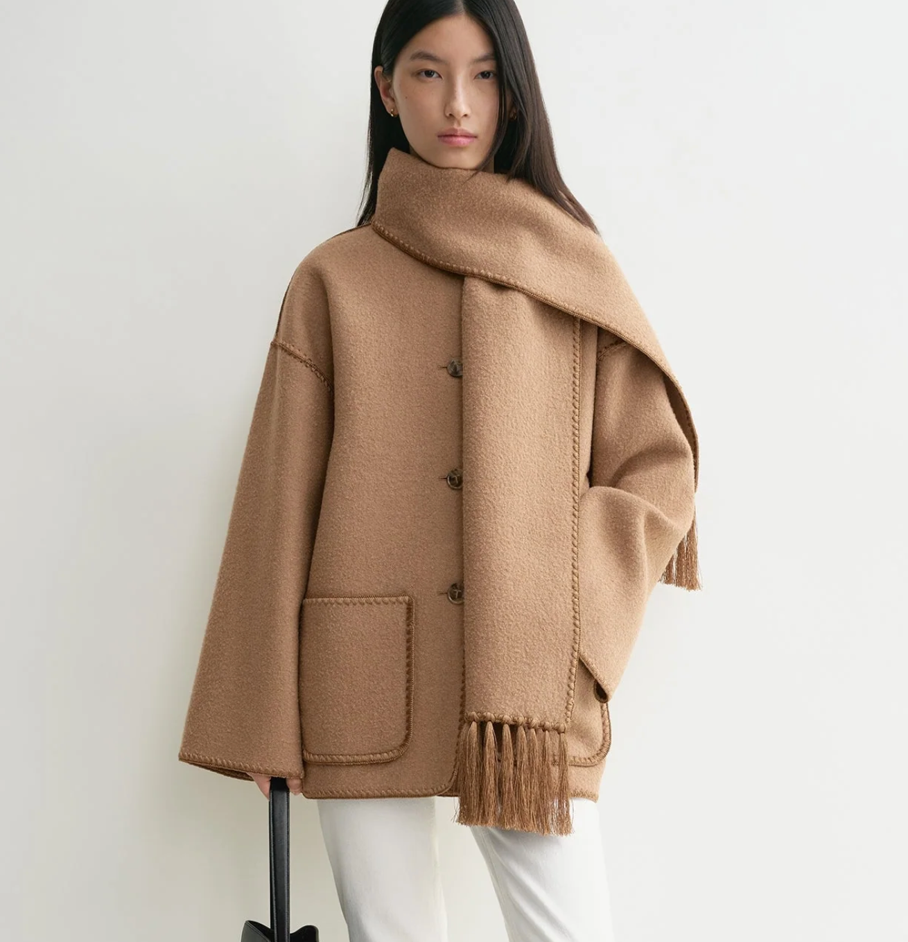 A woman stands against a white background wearing Toteme’s Embroidered Scarf Jacket in camel wool-blend felt with an oversized fit and large pockets, paired with a matching scarf, white pants, and carrying a black handbag.