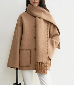 A woman stands against a white background wearing Toteme’s Embroidered Scarf Jacket in camel wool-blend felt with an oversized fit and large pockets, paired with a matching scarf, white pants, and carrying a black handbag.