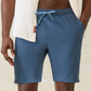 A person stands with one hand in their pocket against a neutral background, wearing a white short-sleeve shirt and Faherty’s Shorelite Swim Trunk in blue, crafted from quick-drying technical fabric.