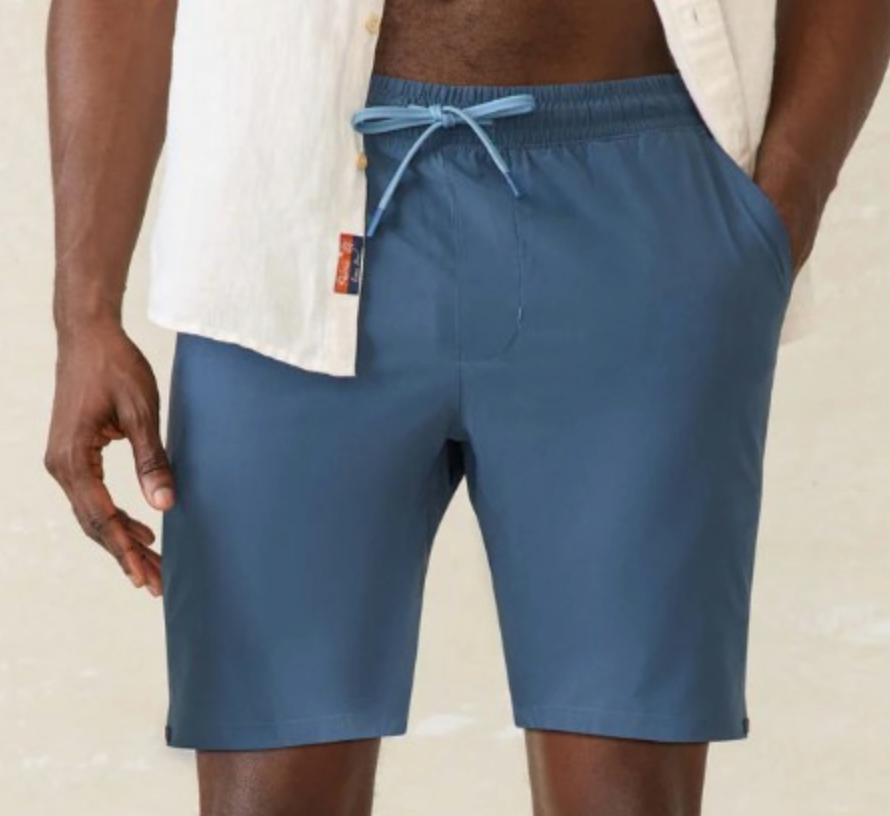 A person stands with one hand in their pocket against a neutral background, wearing a white short-sleeve shirt and Faherty’s Shorelite Swim Trunk in blue, crafted from quick-drying technical fabric.