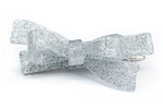 The Bow Glitter Silver Hair Clip by Lillies & Roses features a shimmering silver bow shape with a metallic clasp, evoking the elegance of Brazilian artisan designs, shown against a white background.
