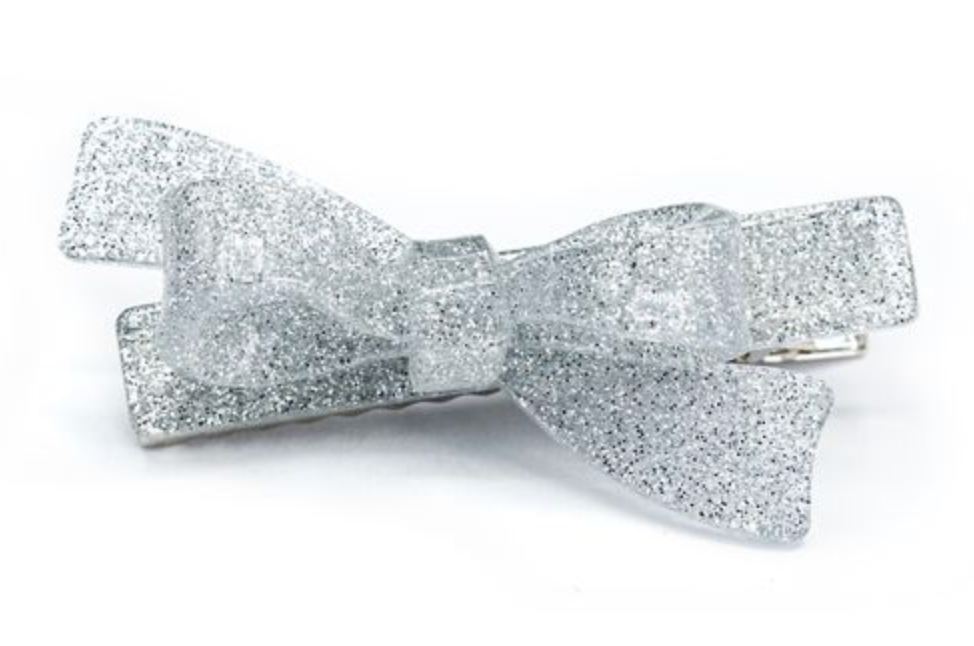 The Bow Glitter Silver Hair Clip by Lillies & Roses features a shimmering silver bow shape with a metallic clasp, evoking the elegance of Brazilian artisan designs, shown against a white background.