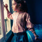 A young girl gazes out a window with one hand on the handle, wearing a light floral blouse and the Louis Louise Girls' Teacher Washed Denim Skirt featuring heart-shaped pockets and an elastic waistband.