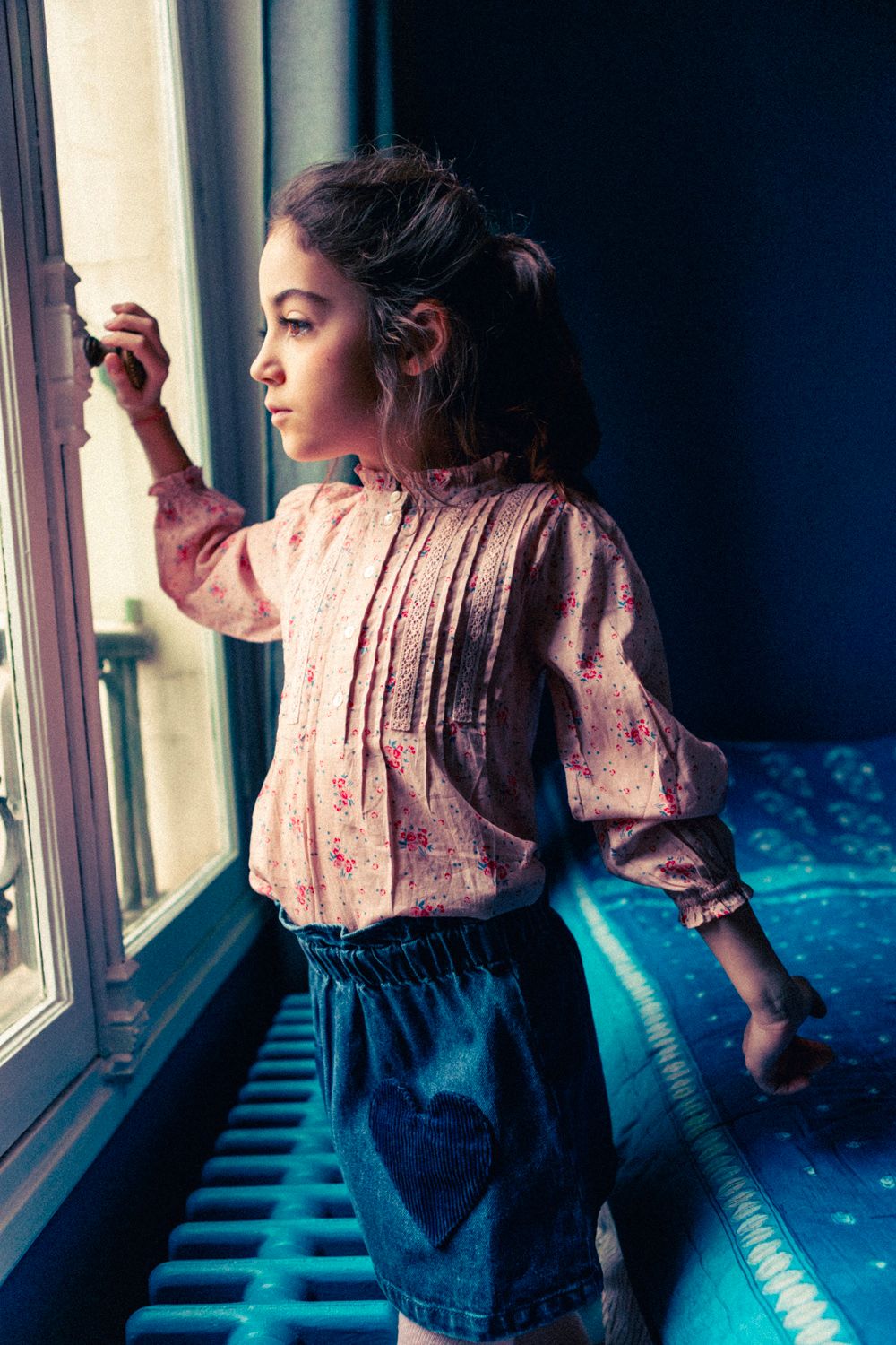 A young girl gazes out a window with one hand on the handle, wearing a light floral blouse and the Louis Louise Girls' Teacher Washed Denim Skirt featuring heart-shaped pockets and an elastic waistband.