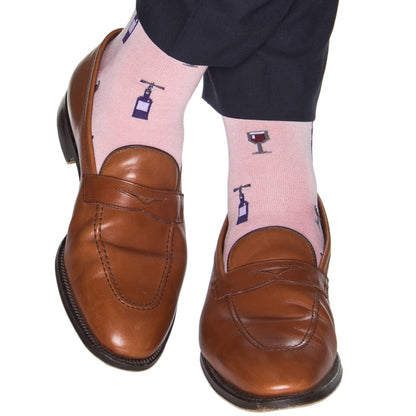 A person wearing tan loafers and the eye-catching Dapper Classics Merlot Mid Calf Socks in pink, adorned with a playful wine glass and bottle design, stands out effortlessly. Crafted with quality mercerized cotton, these stylish socks by Dapper Classics are a testament to fine American craftsmanship, proudly made in the USA.