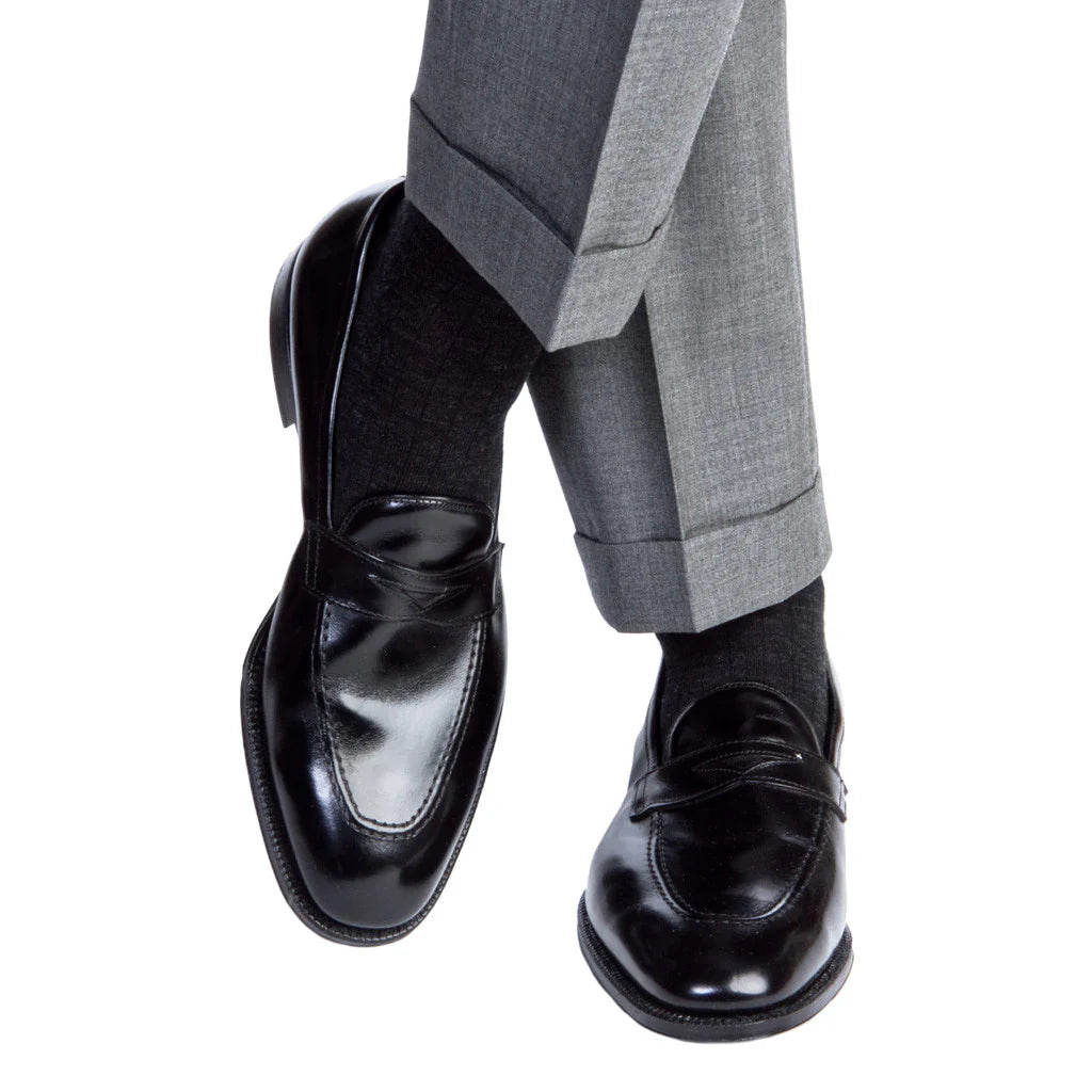 A person wearing Dapper Classics Charcoal Solid Ribbed Fine Merino Wool Sock—mid-calf, linked toe—with black dress shoes and light gray cuffed trousers, one leg crossed over the other against a white background.