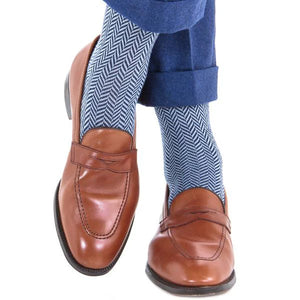 A person wearing Dapper Classics Dress Navy and Sky Blue Herringbone Fine Merino Wool mid-calf socks, cuffed blue trousers, and brown leather loafers sits with legs crossed.