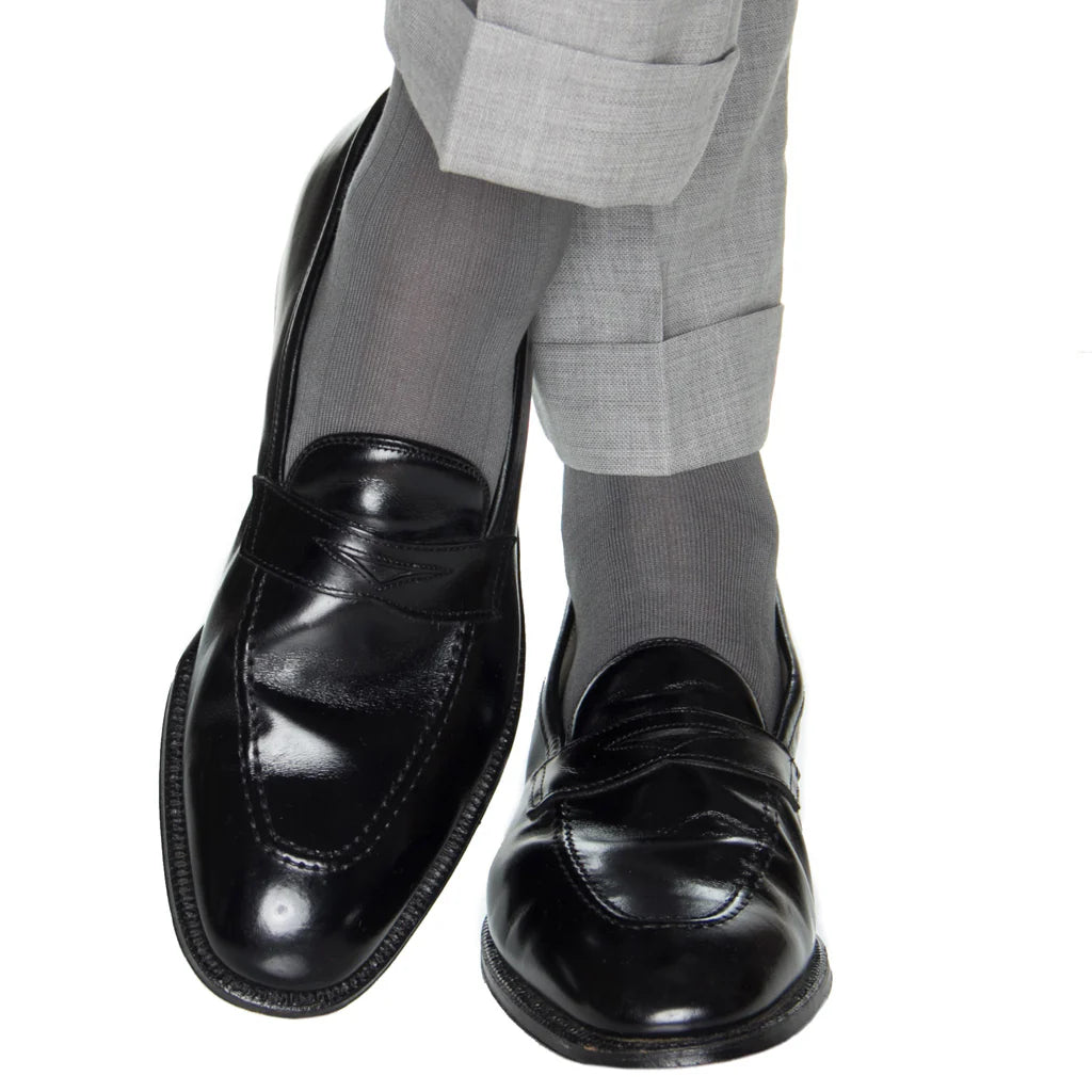 Person wearing light gray dress pants, Dapper Classics Steel Gray Solid Ribbed Cotton Sock Linked Toe Mid-Calf, and black leather loafers, standing with one leg crossed over the other against a white background.