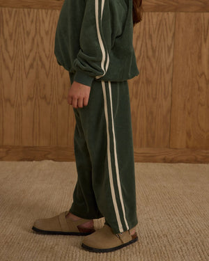 A girl wears Rylee & Cru Baby Girls' and Girls' Track Pant with white side stripes and beige slip-ons, standing on a beige carpet before a wooden wall, highlighting sporty comfort.