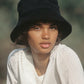 A person wearing the Lack of Color Wave Terry Bucket Hat in black, which has a UPF Rating of 50+, and a white blouse looks directly at the camera outdoors.