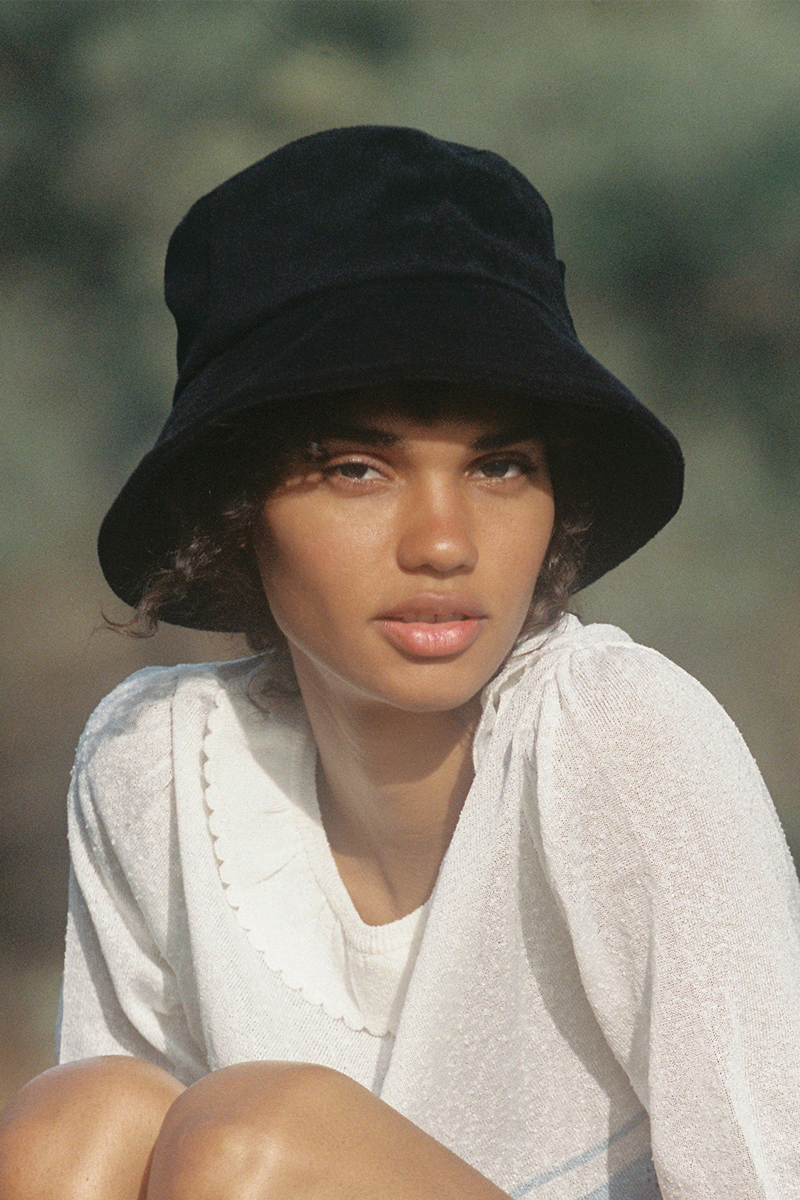 A person wearing the Lack of Color Wave Terry Bucket Hat in black, which has a UPF Rating of 50+, and a white blouse looks directly at the camera outdoors.