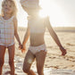 Two kids in swimsuits stand hand in hand, their giggles echoing on the sunny beach. The girl wears Louise Misha's Girls' Primavera Bikini Set with cute floral ruffles, adding whimsy to the scene.