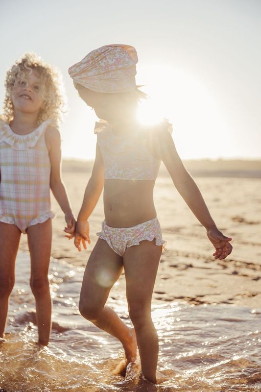 Two kids in swimsuits stand hand in hand, their giggles echoing on the sunny beach. The girl wears Louise Misha's Girls' Primavera Bikini Set with cute floral ruffles, adding whimsy to the scene.