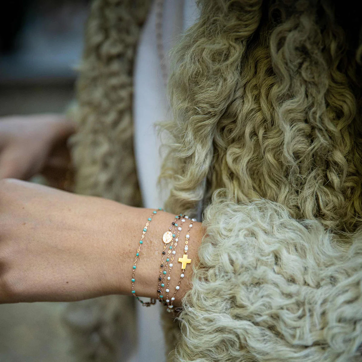 Wearing a fur coat, someone showcases the Gigi Clozeau Classic Cross Charm Bracelet in 18 carat gold, layered with elegant resin pearls.