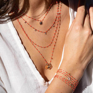 A person adorned in several layers of red beaded necklaces and coordinating bracelets, showcasing a Gigi Clozeau Lucky Heart Pendant crafted in 18-carat gold, styled with a white top.