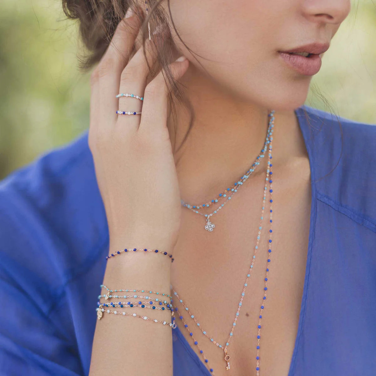 A woman in a blue top, adorned with layered silver and blue necklaces, bracelets, rings, and the Gigi Clozeau Diamond Lucky Clover Pendant, touches her face delicately.