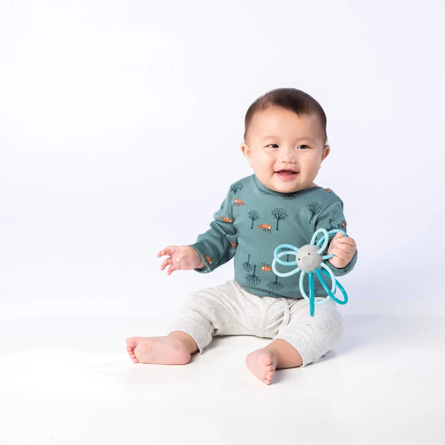 A baby wearing a green shirt with animal prints and gray pants sits on the floor holding a Manhattan Toy Winkel Elephant teething toy.