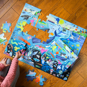 A child assembles the Eeboo Within the Sea 48 Piece Puzzle on a wooden floor, surrounded by vibrant aquatic creatures, making playtime fun and educational with several large pieces still to fit.